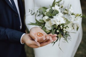 Trouver le bon prestataire mariage est la clé d’un mariage réussi. Découvrez nos conseils pour bien choisir, collaborer en confiance et vivre votre journée sans stress. Anneaux de mariage dans les mains des mariés et avec un beau bouquet de mariée composé de verdure et de fleurs blanches